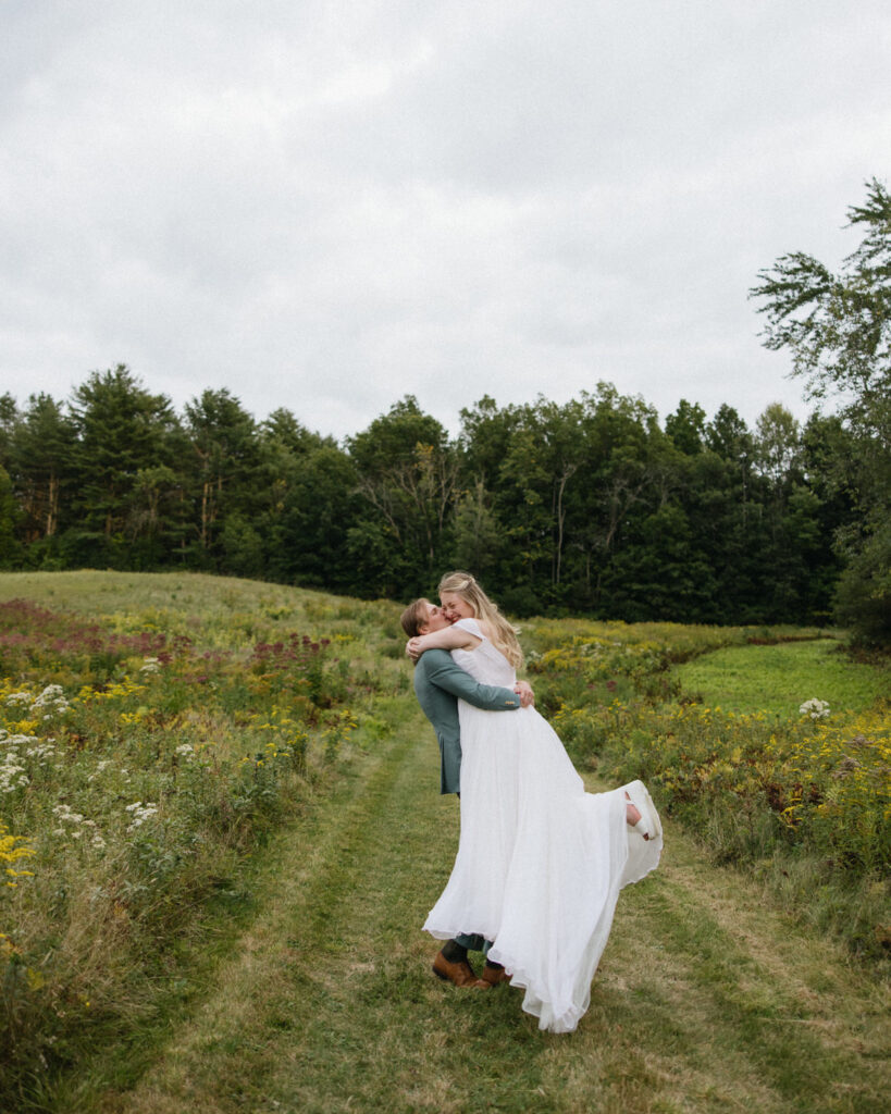 Groom lifts up bride in meadow of wedding venue in Maine